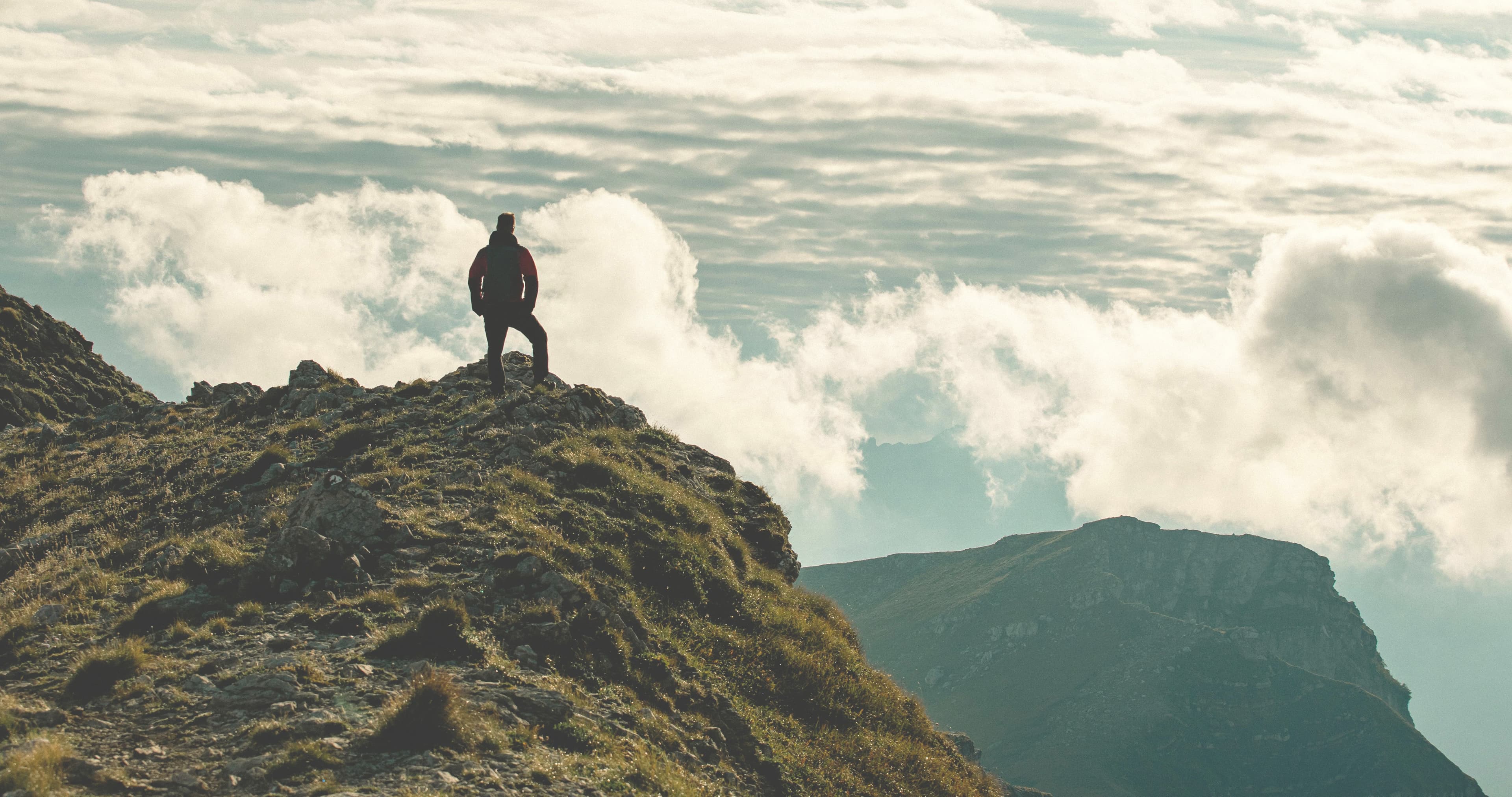 A silhouette on the top of a mountain in the foreground with clouds shown in the background.