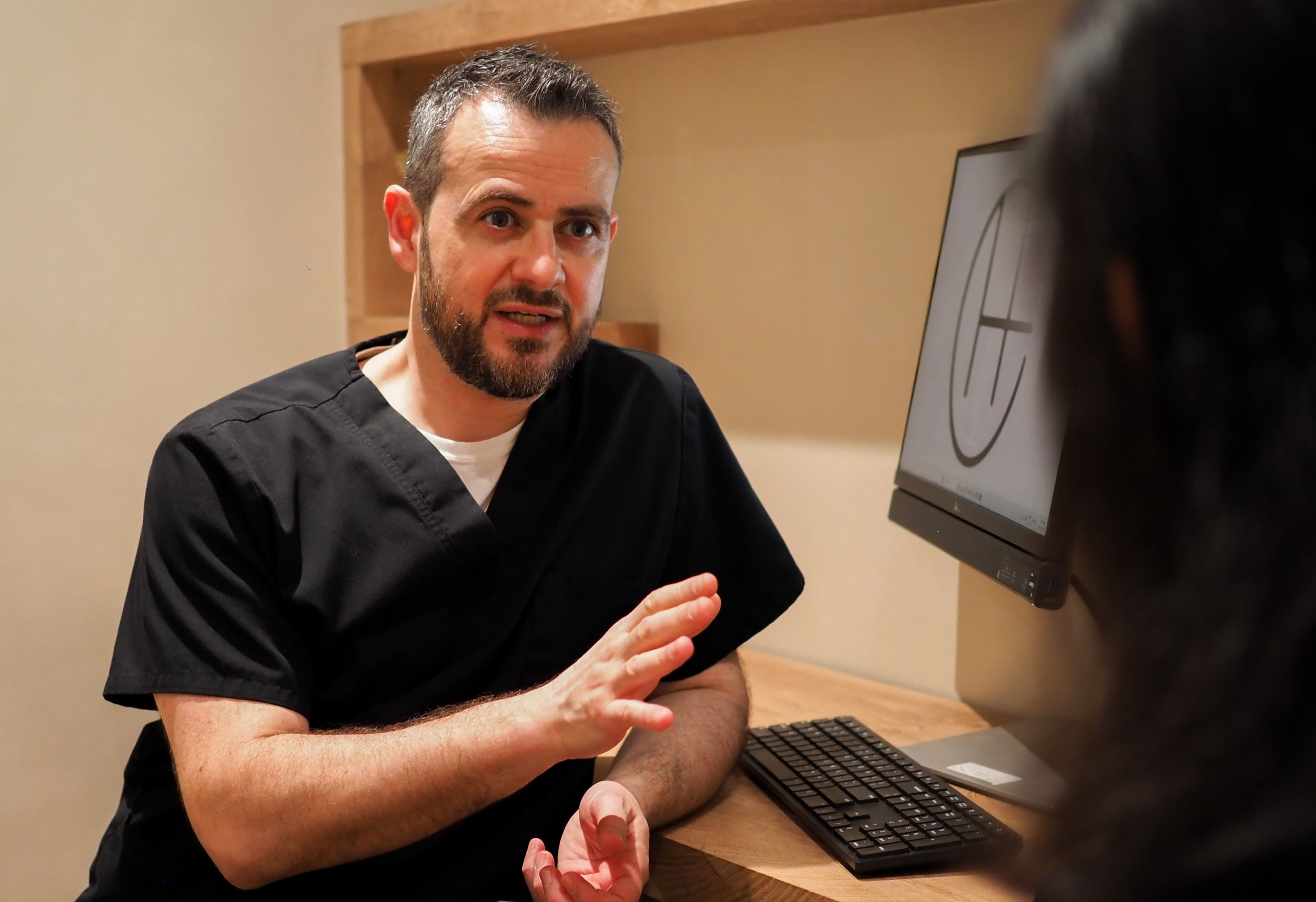 Dr. David Gabriele in scrubs speaking with a patient at his desk beside a monitor.