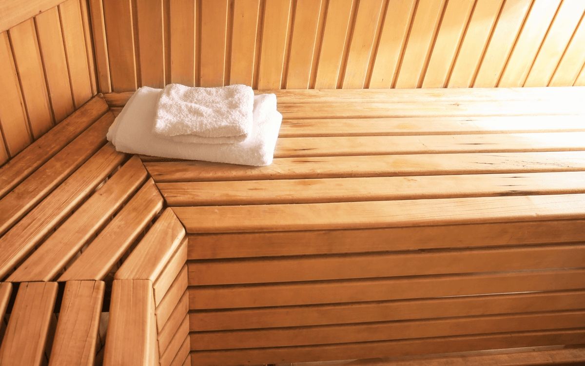 Woman in white swimsuit relaxing on wooden bench inside a sauna, leaning back with eyes closed in a peaceful pose"