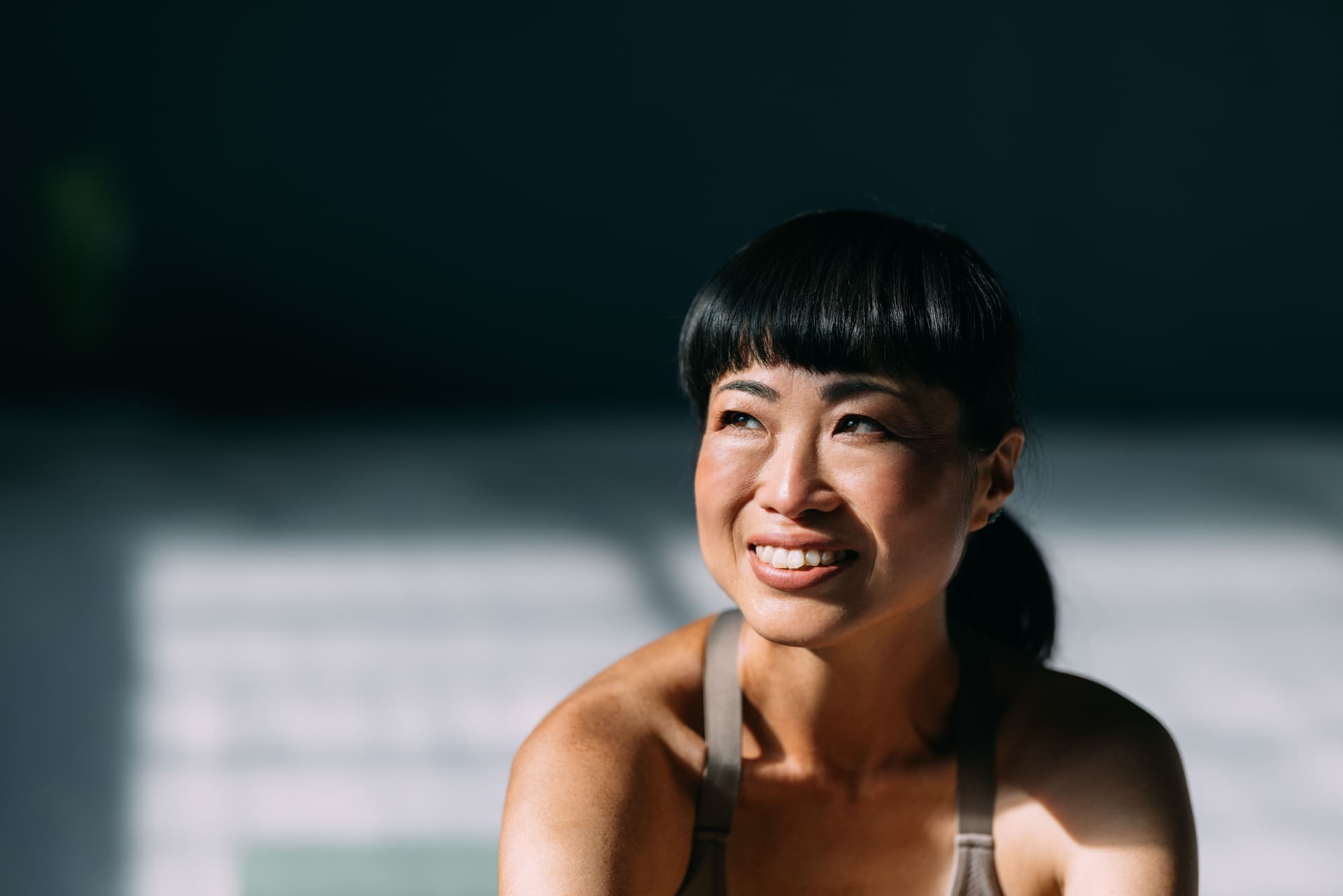 Portrait photograph of a smiling woman with bangs in athletic wear, captured in natural lighting with dramatic shadows.