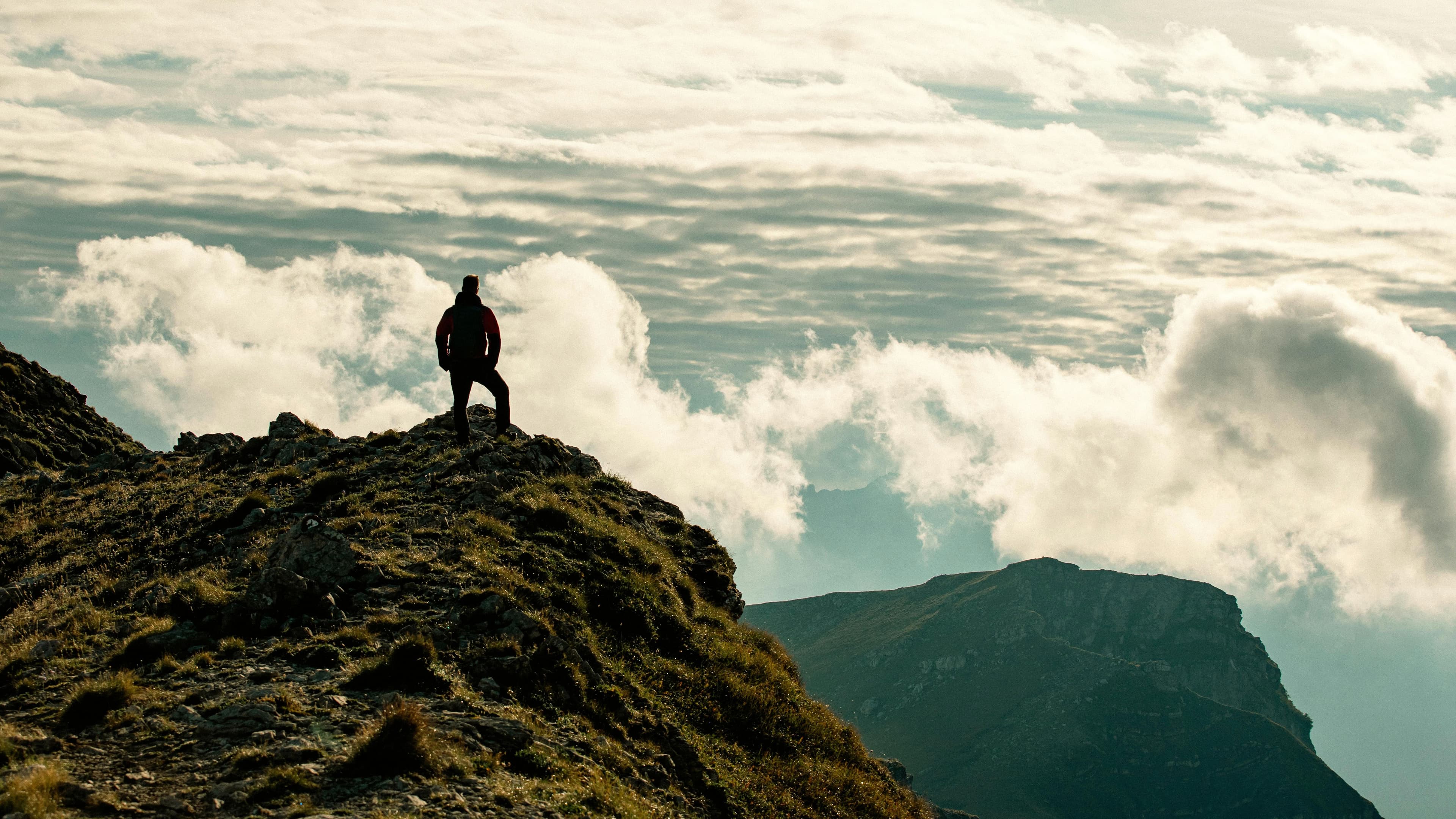 A silhouette on the top of a mountain in the foreground with clouds shown in the background.