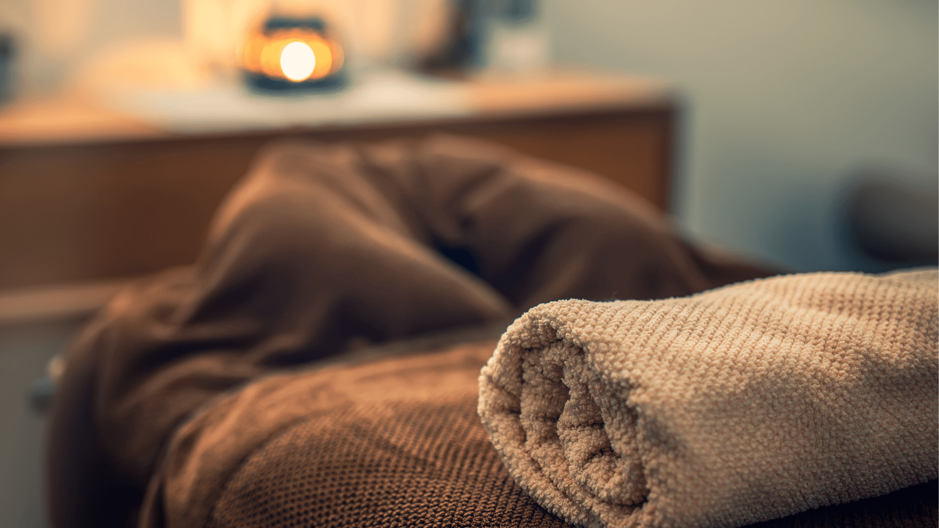 Photo of spa setting with rolled towel in foreground and candlelight in background, creating a relaxing atmosphere.