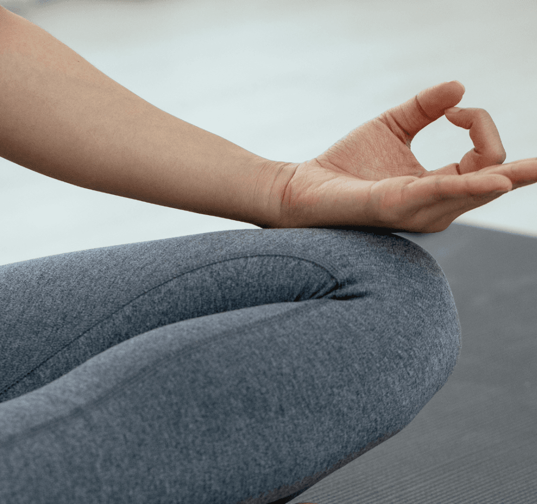 Close-up photo of a person sitting cross-legged in a meditative pose on a yoga mat, with one hand in a mudra gesture.