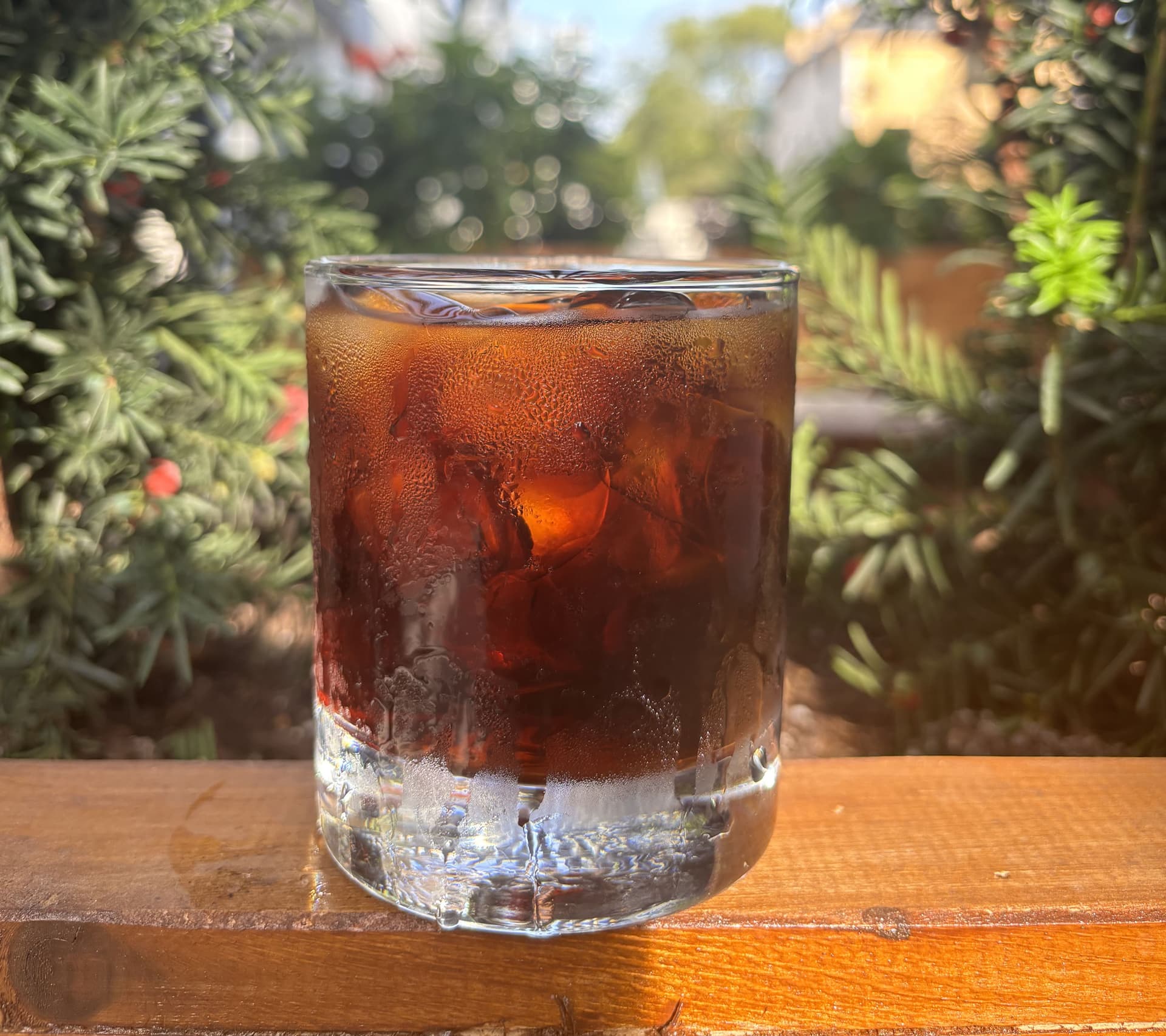 Here's the alt text: Alt Text: "Textured glass of iced coffee on a wooden table with lush greenery and a pergola visible in the blurred background
