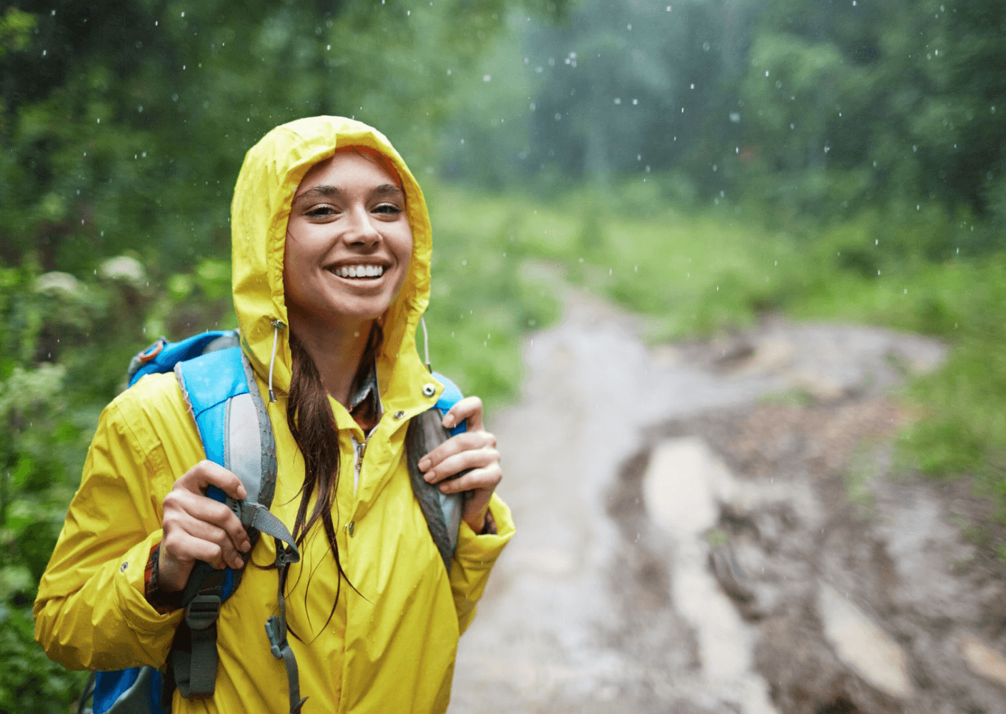 Photo of a smiling woman in a yellow raincoat and backpack hiking through a rainy forest trail.