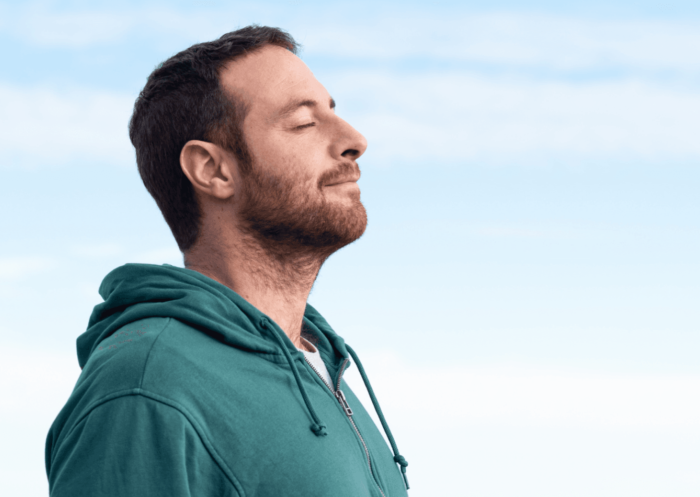 Photo of a man in a green hoodie enjoying a moment of mindfulness outdoors with closed eyes and a calm expression.