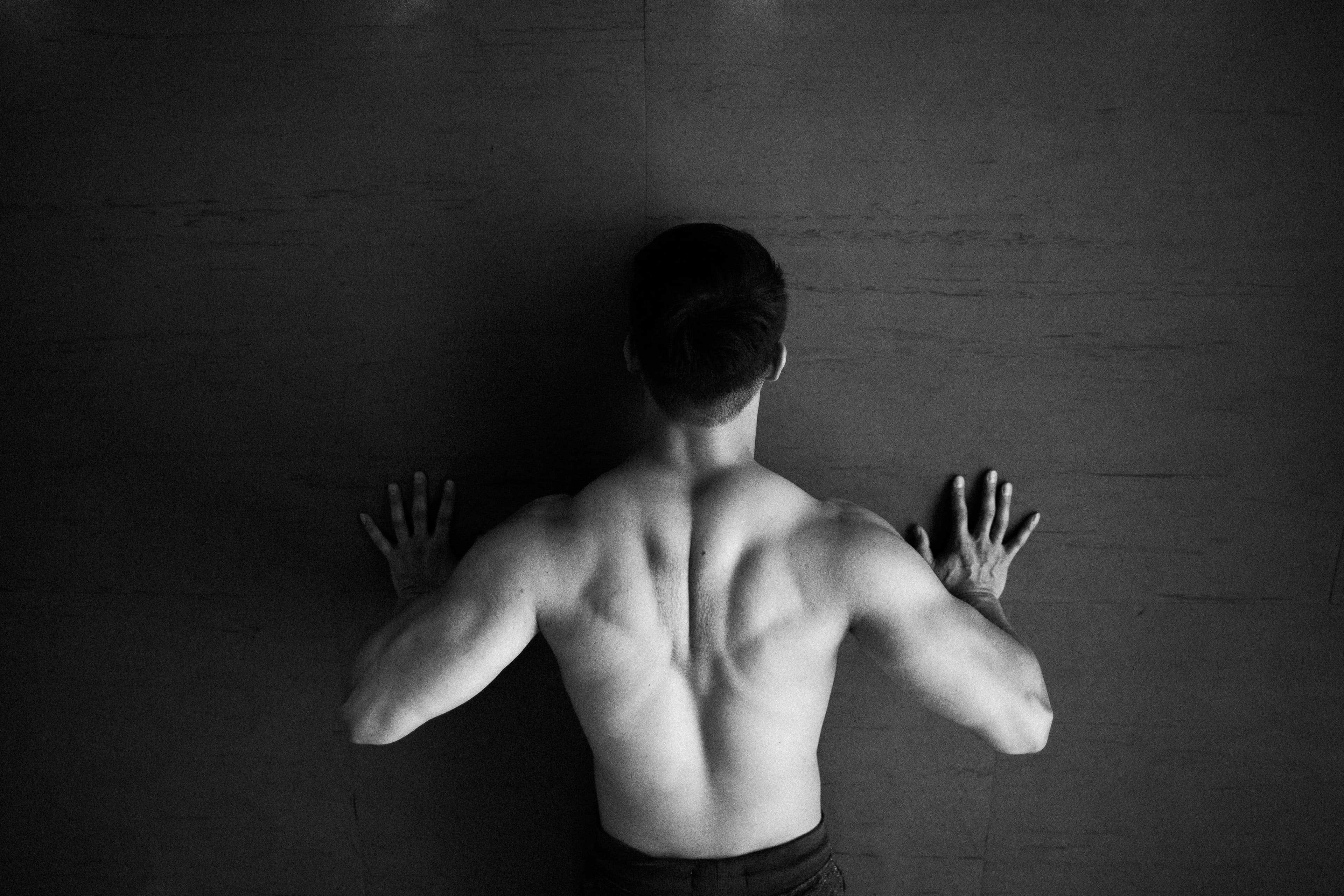 Black and white photograph showing a muscular bare back against the ground, hands pressed outward, suggesting strength and vulnerability. Artistic portrait with dramatic lighting and shadow.