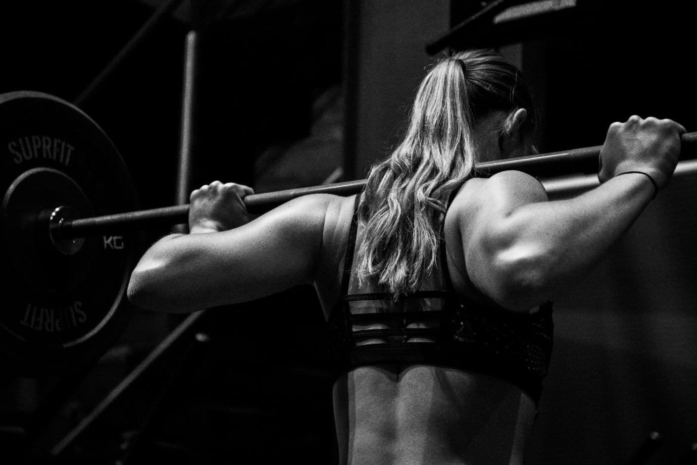 A woman holding a barbell across her shoulders, photographed from behind.