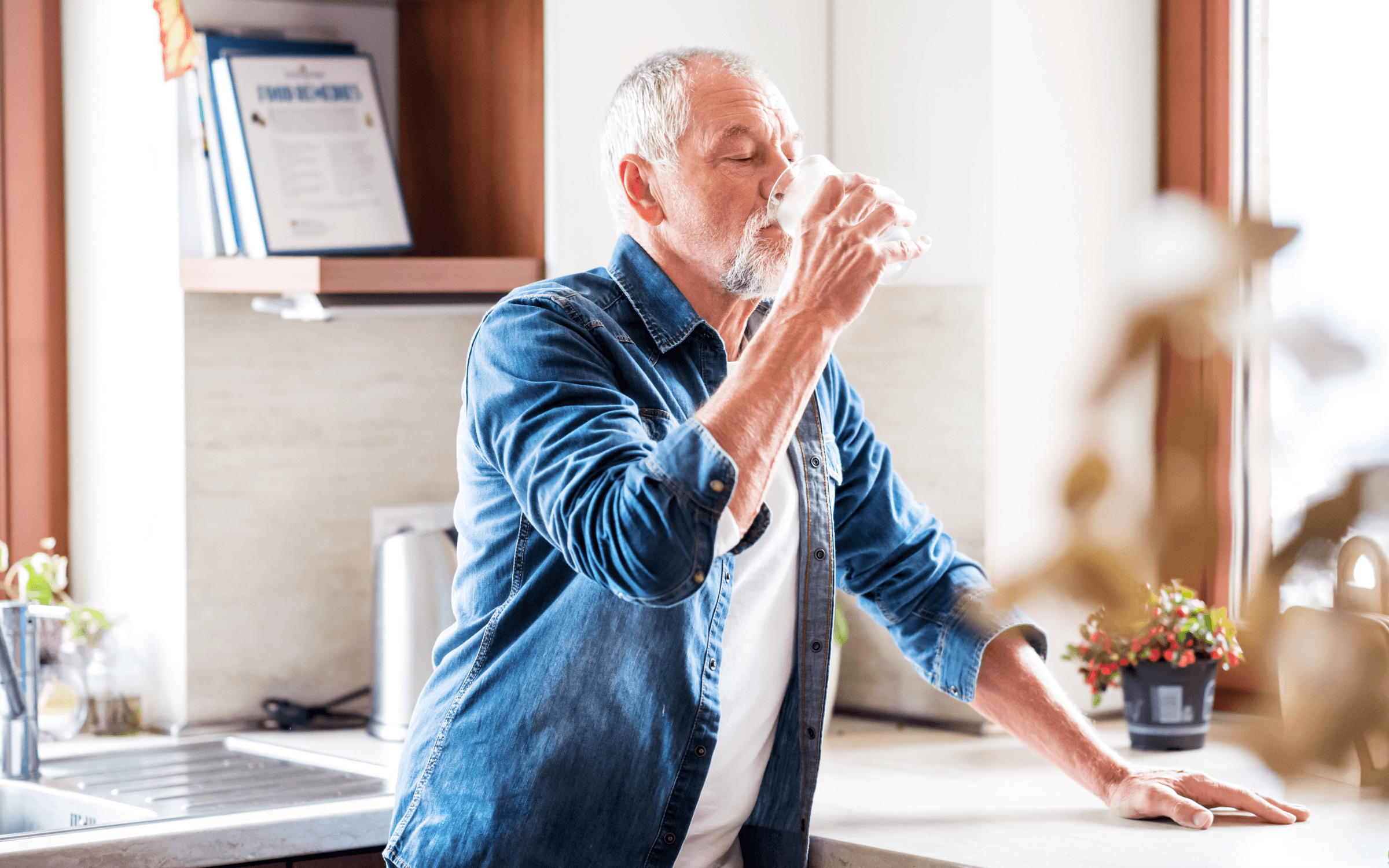 An older man drinking a glass of water in a kitchen.